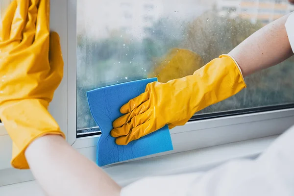 Man in yellow rubber gloves cleaning window glasse with cleaner spray detergent and squeegee or rag at home or office, copy space. Housework and housekeeping, home hygiene. Blue sky view, plastic window frames