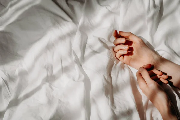 Elegant hands lie on the white bed sheet in the sunlight. Bed with white linens. The concept of a good morning, stress relief, self-care, relaxation and time for yourself