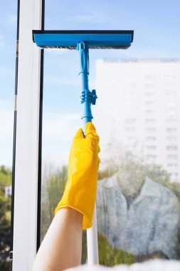 Man in yellow gloves cleaning window with squeegee and spray detergent at home terrace. House cleaning and house chores, domestic hygiene. Window cleaning background with blue sky