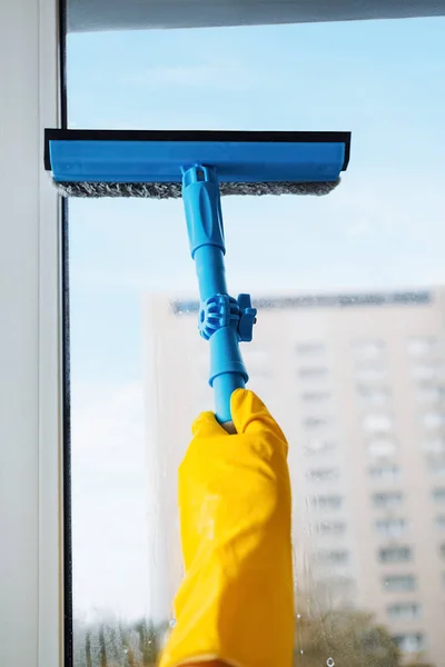 Man in yellow gloves cleaning window with squeegee and spray detergent at home terrace. House cleaning and house chores, domestic hygiene. Window cleaning background with blue sky