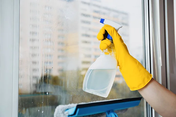 Man in yellow gloves cleaning window with squeegee and spray detergent at home terrace. House cleaning and house chores, domestic hygiene. Window cleaning background with blue sky