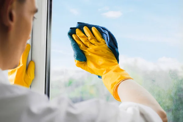 Man in yellow gloves cleaning window with rag and spray detergent at home terrace. House cleaning and house chores, domestic hygiene. Window cleaning background with blue sky