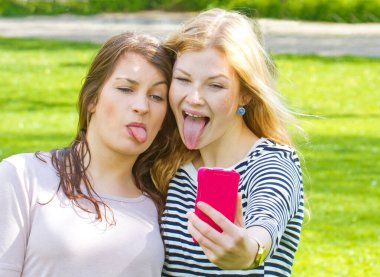 İki kız akıllı telefonlarıyla selfie çekiyor ve dillerini dışarı çıkarıyor.