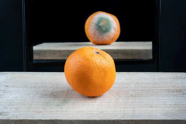 concept of seeing the real face of something in the mirror. Fresh and ripe orange fruit on table against mirror reflection with rotten bad side. Fruit with selective focus.