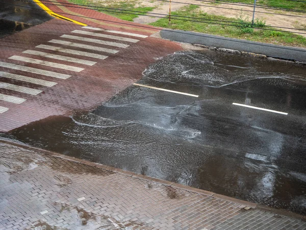 Heavy rain drops falling on city street during downpour, pedestrian crossing and the road with puddles