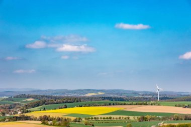 İlkbaharda güzel kırsal alanlar, çiçek açan kolza tohumu tarlaları, buğday yeşil tarlaları, uzaktaki rüzgar santrali, beyaz bulutlu mavi gökyüzü, Kuzey Ren-Vestfalya, Almanya