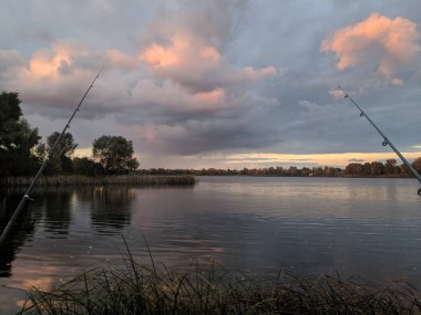 Nehirde balık tutmak. Elinde olta olan bir balıkçı. Yakalayın. Olta kamışı tekeri yakın çekim, gün doğumuyla balık tutma. Elinde olta ve kova olan küçük bir kız balık tutmaya geldi ve gölete baktı..