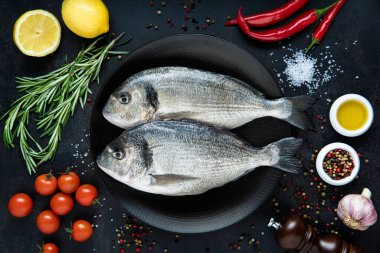 Siyah bir tabakta iki dorado balığı, baharat, biber, biberiye, domates ve koyu arkaplanda limon. Üst görünüm