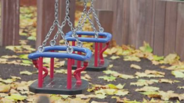 Empty toddler swing, empty and swaying in breeze with autumn leaves on the ground.