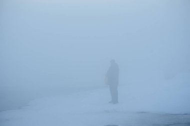 Kuzey buzlu manzarası. Dondurulmamış bir nehrin yüzeyinden sis yükseliyor. Siste, ördekleri beslemek için nehir kenarına gelen bir adamın silueti görünür..