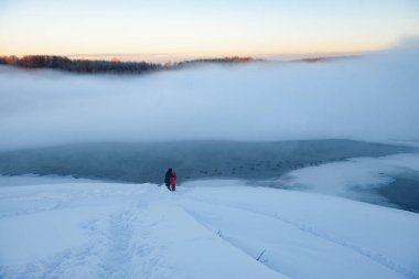 Kuzey buzlu manzarası. Dondurulmamış bir nehrin yüzeyinden sis yükseliyor. Anne ve kızı nehir kıyısında durup suda yüzen ördeklere bakıyorlar..