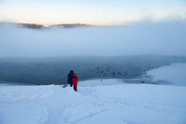 Kuzey buzlu manzarası. Dondurulmamış bir nehrin yüzeyinden sis yükseliyor. Anne ve kızı nehir kıyısında durup suda yüzen ördeklere bakıyorlar..
