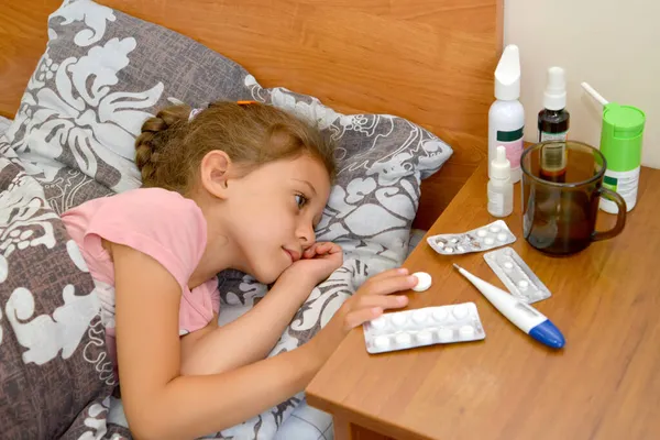 A five-year-old girl lies in bed and takes a pill with her hand. Self-medication