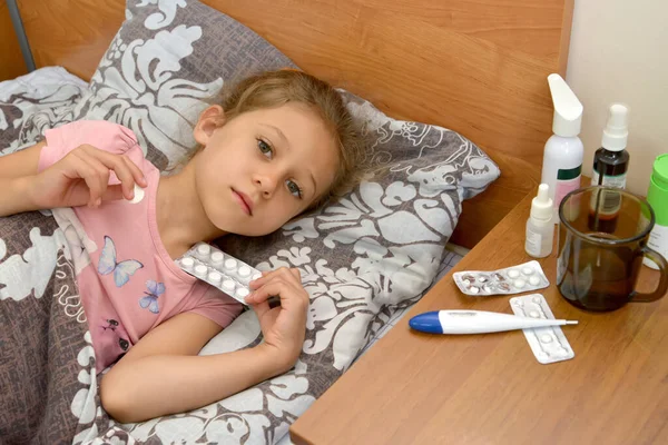 A sick girl lies in bed and holds pills in her hands. Self-medication