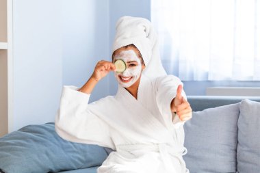 Elinde salatalık dilimleri olan yüz maskeli genç bir kadın. Yüzünde yüz maskesi olan genç ve güzel bir kadın elinde taze salatalık dilimleri tutuyor. Cilt bakımı ve tedavisi, spa, doğal güzellik ve kozmetoloji konsepti