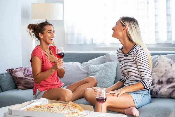 Happy young female friends eating pizza with wine on sofa at home. Happy smiling young women friends eating pizza and opening boottle of wine at home