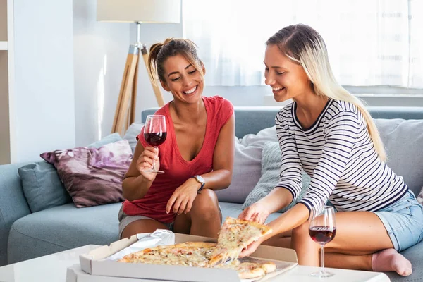 Happy young female friends eating pizza with wine on sofa at home. Happy smiling young women friends eating pizza and opening boottle of wine at home