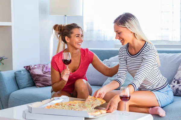Happy young female friends eating pizza with wine on sofa at home. Happy smiling young women friends eating pizza and opening boottle of wine at home. Girls havin fun