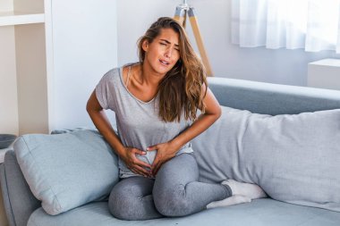 Kanepede hasta bir şekilde yatan bir kadın. Güzel genç bir kadın yatağa uzanıyor ve ellerini karnının üstüne koyuyor. Yatakta karın ağrısı çeken bir kadın. Adet dönemi.