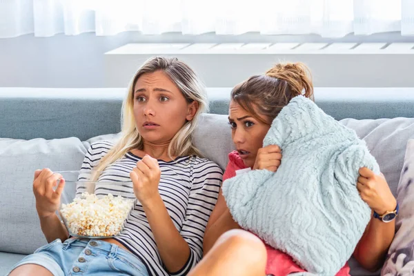 Terrified friends watching tv in the night sitting on a couch in the living room at home. Scary Movie Night with Girlfriends, Frightened young women watching scary movie at home.