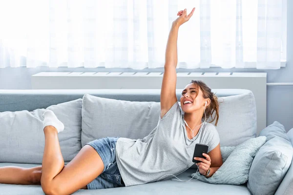 Happy woman or teenage girl in headphones listening to music from smartphone and dancing on bed at home. Young woman listening to music online on smartphone in headphones