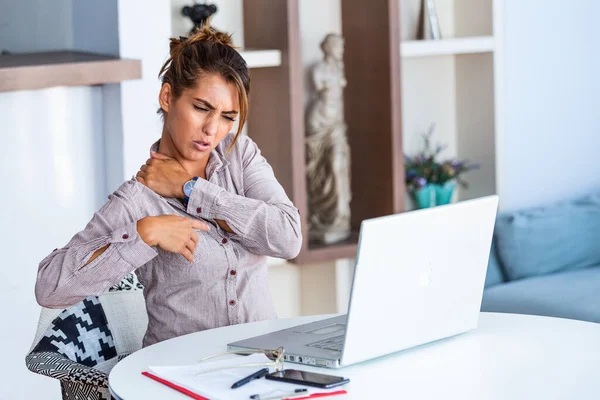 Portrait of young stressed woman sitting at home office desk in front of laptop, touching aching back with pained expression, suffering from backache after working on pc