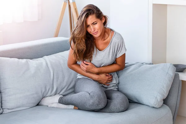 Kanepede hasta bir şekilde yatan bir kadın. Güzel genç bir kadın yatağa uzanıyor ve ellerini karnının üstüne koyuyor. Yatakta karın ağrısı çeken bir kadın. Adet dönemi.