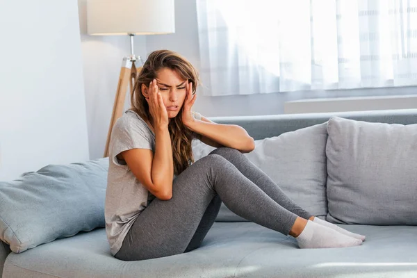 Portrait of an attractive woman sitting on a sofa at home with a headache, feeling pain and with an expression of being unwell. Upset depressed woman lying on couch feeling strong headache migraine.