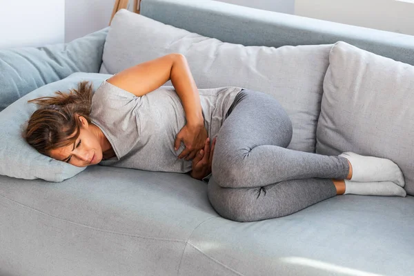 Adet gören karın ağrısına karşı el ele tutuşan, evde üzgün yatan, kadın sağlığı konseptinde mide krampı yaşayan bir kadın.