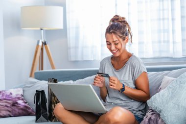 Kredi kartı tutan ve dizüstü bilgisayar kullanan genç bir kadın. İnternetten alışveriş konsepti. Mutlu kadın evde online alışveriş yapıyor.