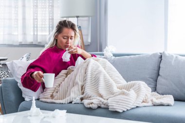 Hasta kadın. Flu. kadın üşüttü. Dokuların içine hapşırmak. Baş ağrısı. Virüs. İlaçlar. Burnunu mendile sümküren soğuk algınlığına yakalanmış genç bir kadın. Kanepede oturan, başı ağrıyan hasta kadın.