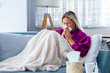 Hasta kadın. Flu. kadın üşüttü. Dokuların içine hapşırmak. Baş ağrısı. Virüs. İlaçlar. Burnunu mendile sümküren soğuk algınlığına yakalanmış genç bir kadın. Kanepede oturan, başı ağrıyan hasta kadın.