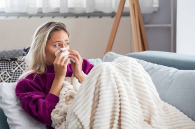 Hasta kadın. Flu. kadın üşüttü. Dokuların içine hapşırmak. Baş ağrısı. Virüs. İlaçlar. Burnunu mendile sümküren soğuk algınlığına yakalanmış genç bir kadın. Kanepede oturan, başı ağrıyan hasta kadın.