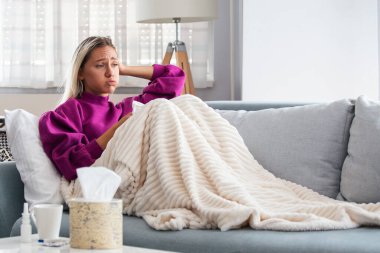 Hasta kadın. Flu. kadın üşüttü. Dokuların içine hapşırmak. Baş ağrısı. Virüs. İlaçlar. Burnunu mendile sümküren soğuk algınlığına yakalanmış genç bir kadın. Kanepede oturan, başı ağrıyan hasta kadın.