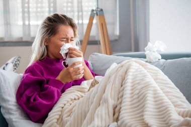 Hasta kadın. Flu. kadın üşüttü. Dokuların içine hapşırmak. Baş ağrısı. Virüs. İlaçlar. Burnunu mendile sümküren soğuk algınlığına yakalanmış genç bir kadın. Kanepede oturan, başı ağrıyan hasta kadın.
