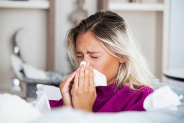 Hasta kadın. Flu. kadın üşüttü. Dokuların içine hapşırmak. Baş ağrısı. Virüs. İlaçlar. Burnunu mendile sümküren soğuk algınlığına yakalanmış genç bir kadın. Kanepede oturan, başı ağrıyan hasta kadın.