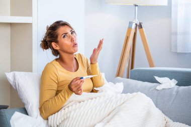 Hastalık, mevsimsel virüs problemi konsepti. Kanepede uzanmış, termometredeki sıcaklığa bakarak hasta olan bir kadın. Hasta kadın yüksek ateşle yatakta yatıyor. Soğuk algınlığı ve migren.