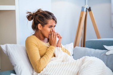 Hasta kadın. Flu. kadın üşüttü. Dokuların içine hapşırmak. Baş ağrısı. Virüs. İlaçlar. Burnunu mendile sümküren soğuk algınlığına yakalanmış genç bir kadın. Kanepede oturan, başı ağrıyan hasta kadın.