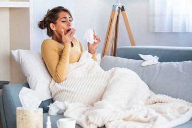 Hasta kadın. Flu. kadın üşüttü. Dokuların içine hapşırmak. Baş ağrısı. Virüs. İlaçlar. Burnunu mendile sümküren soğuk algınlığına yakalanmış genç bir kadın. Kanepede oturan, başı ağrıyan hasta kadın.
