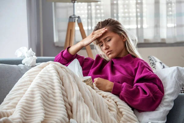 Sick woman with headache sitting under the blanket. Sick woman with seasonal infections, flu, allergy lying in bed. Sick woman covered with a blanket lying in bed with high fever and a flu, resting.