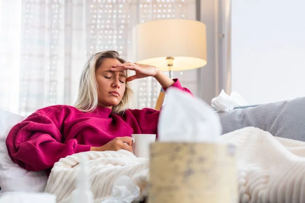 Sick woman with headache sitting under the blanket. Sick woman with seasonal infections, flu, allergy lying in bed. Sick woman covered with a blanket lying in bed with high fever, Coronavirus symptoms