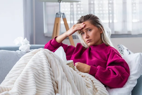 Sick woman with headache sitting under the blanket. Sick woman with seasonal infections, flu, allergy lying in bed. Sick woman covered with a blanket lying in bed with high fever and a flu, resting.