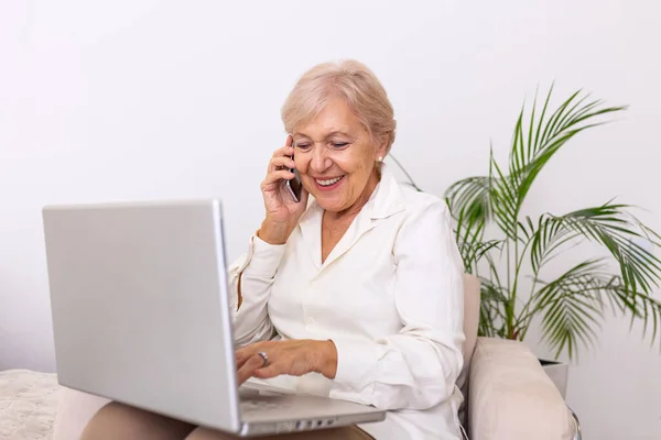 Elderly woman working on laptop computer, smiling, talking on the phone. Senior woman using laptop. Elderly woman sitting at home, using laptop computer and talking on her mobile phone, smiling.