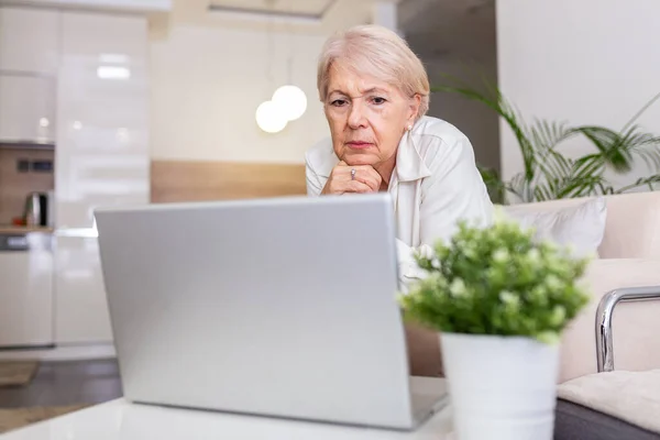 Portrait of senior woman sitting at table at home and working on her laptop. Older lady surfing the net from home while sitting on her sofa and using laptop computer