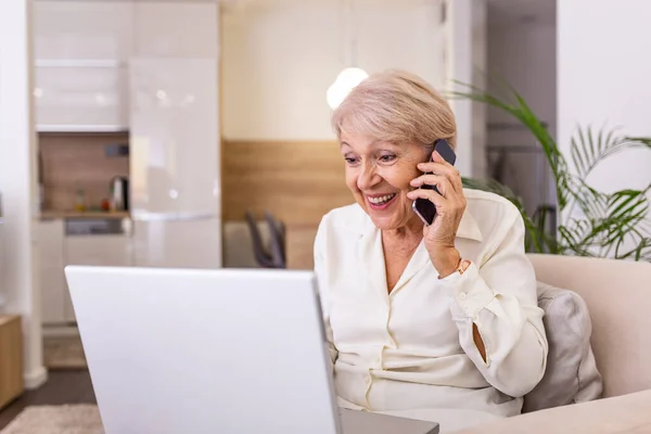 Elderly woman working on laptop computer, smiling, talking on the phone. Senior woman using laptop. Elderly woman sitting at home, using laptop computer and talking on her mobile phone, smiling.