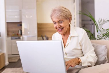 Laptopla çalışan yaşlı bir bayan. Evde dizüstü bilgisayarda çalışan yaşlı güzel bir kadının portresi. Üst düzey kadın evde dizüstü bilgisayar kullanıyor, gülüyor.