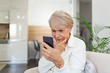 Evde cep telefonu kullanmakta zorlanan kafası karışmış yaşlı bir kadın. Kanepede beyaz saçlı yaşlı bir kadın oturuyor ve akıllı telefonuyla mesajlaşmaya çalışıyor..