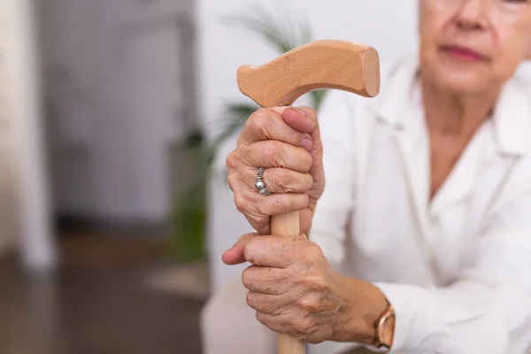 Hands of an old woman with a cane, Elder lady sitting on the couch with wooden walking stick. Cropped shot of a senior woman holding a cane in a retirement home