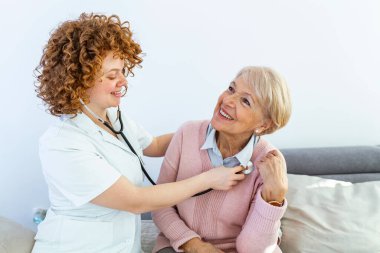 Ev ziyareti sırasında sağlık ziyaretçisi ve yaşlı bir kadın. Genç bayan doktor kıdemli hastayı muayene ediyor. Beyaz önlüklü genç bayan doktor kıdemli kadını muayene ediyor..