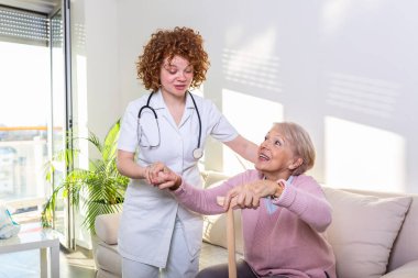 Yaşlı bir kadının oturma odasındaki kanepeden kalkmasına yardım eden bir kadın bakıcı. Gülümseyen hemşire, yaşlı kadına kalkması için yardım ediyor. Kanepeden kalkarken hastaya bakan hemşire.